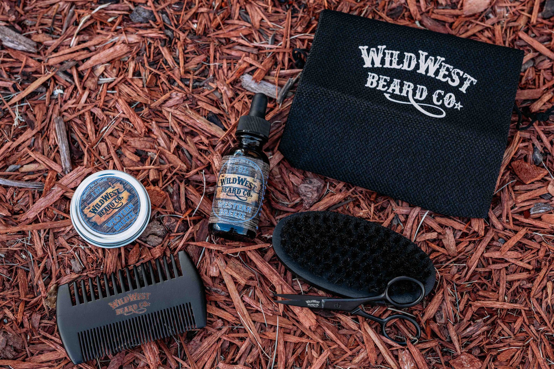 A clean picture of the Wild West Beard Co beard care starter kit contents on a nice red mulch background.  With a travel bag, beard comb, beard brush, natural beard oil, natural beard balm, and snipping scissors.